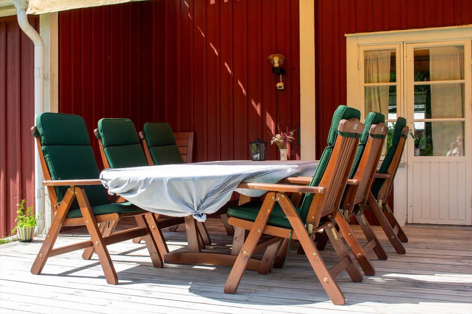 Holztisch mit sechs grünen Stühlen auf einer Terrasse vor einem roten Gebäude.