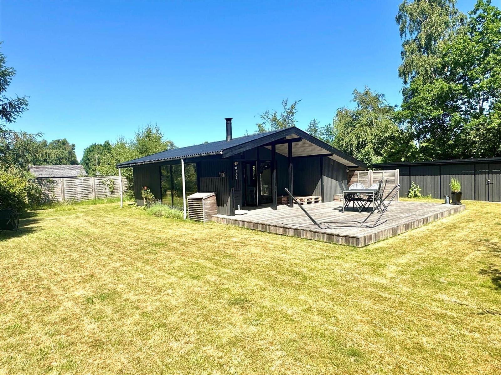 Moderne Hütte mit Terrasse, Garten und Grillplatz. Umgeben von Bäumen und Zaun.
