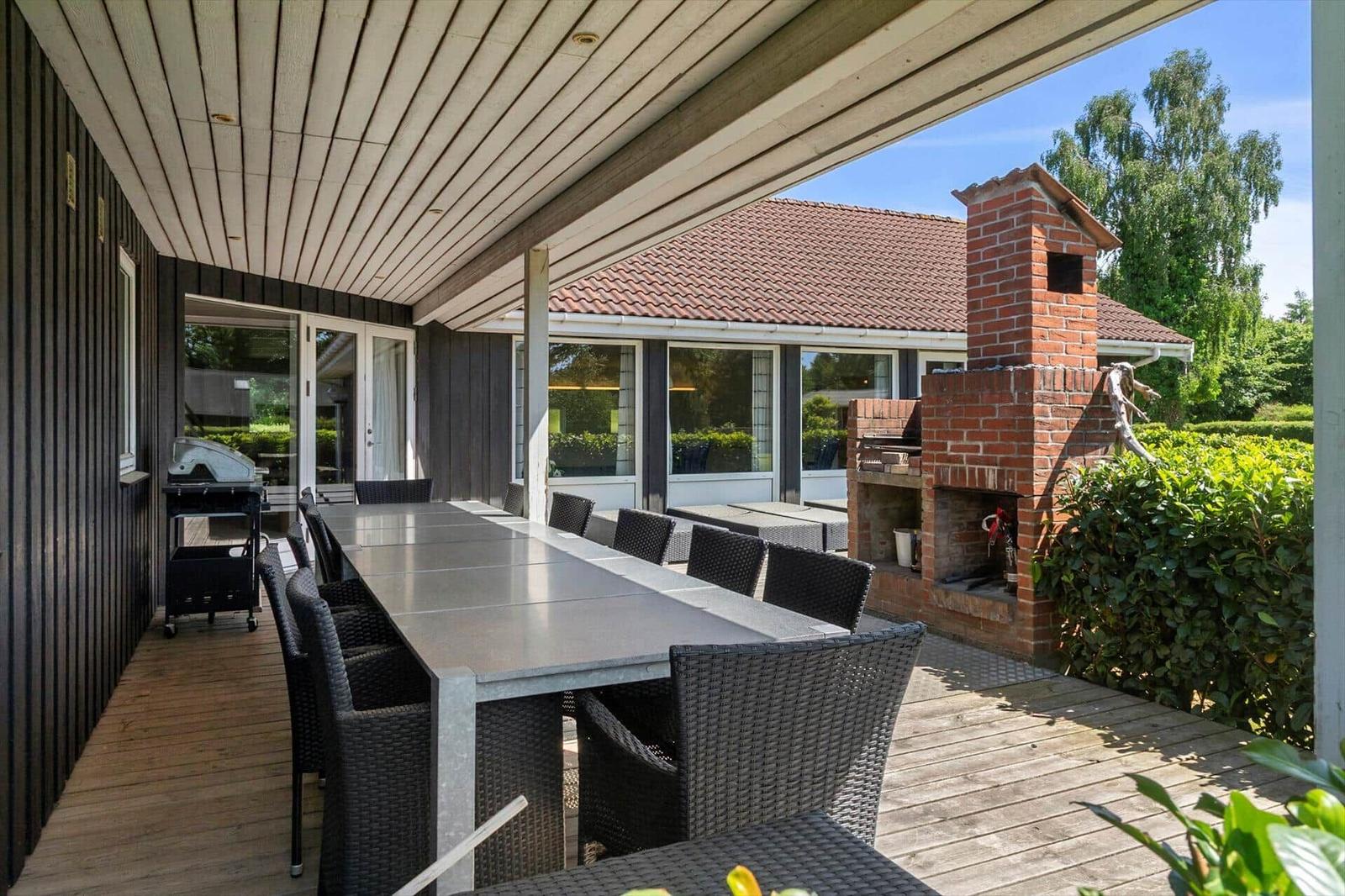 Terrasse mit Tisch, Stühlen, Grill und Backofen. Hintergrund: Haus und Bäume.