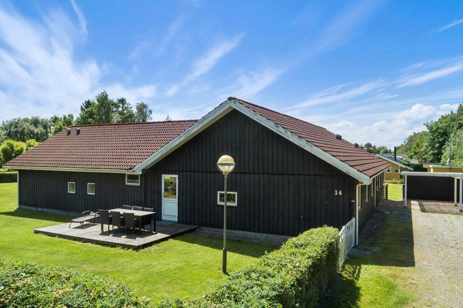 Ein dunkles Holzhaus mit Terrasse, Garten und rotem Dach unter blauem Himmel.