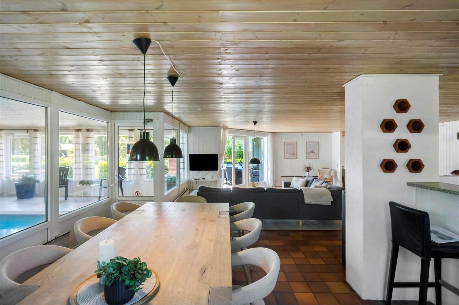 Essbereich mit langem Tisch, Sofa im Hintergrund und Poolbereich durch Fenster sichtbar.