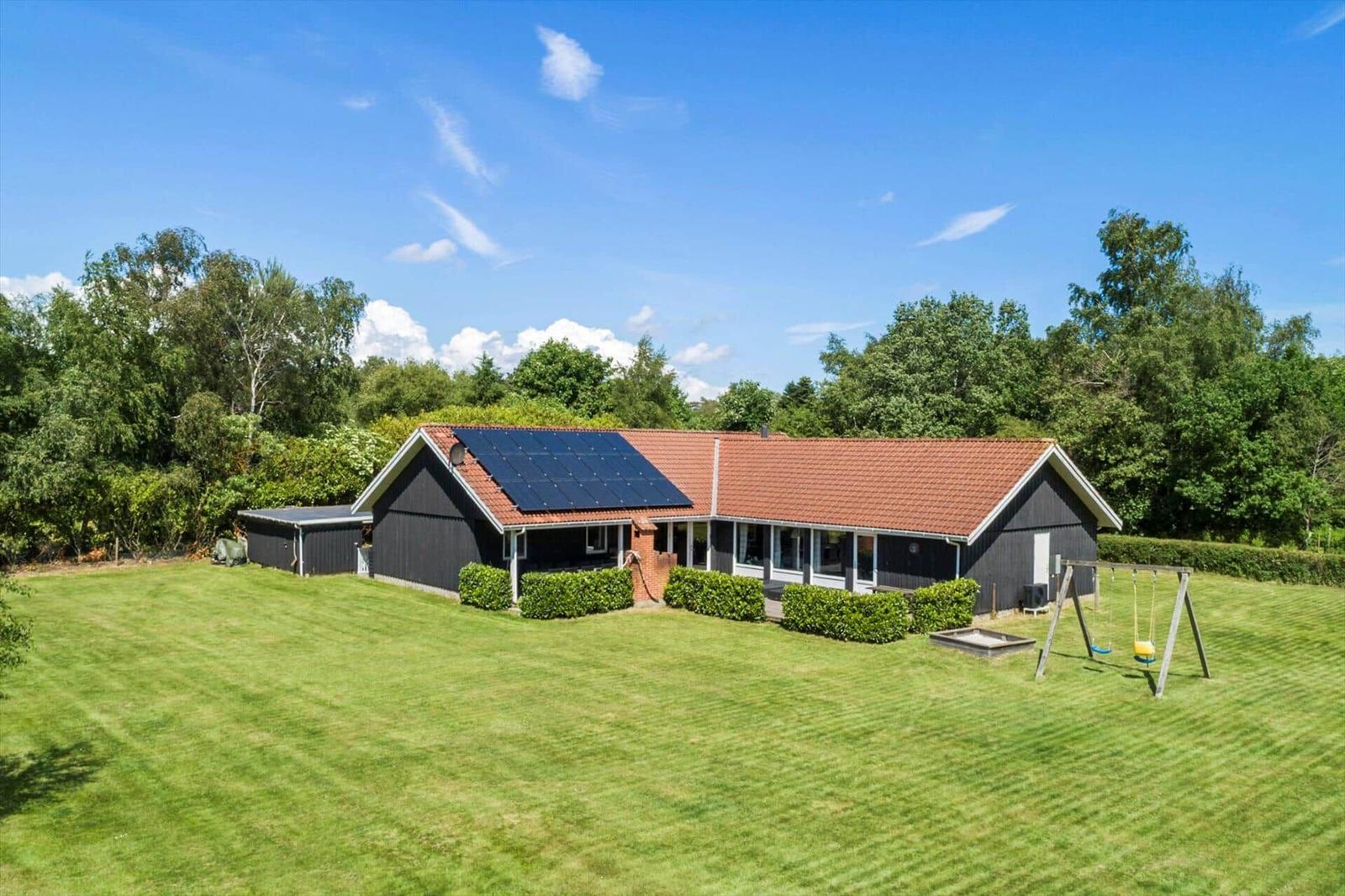 Haus mit Solaranlage, Garten und Schaukel. Umgeben von Bäumen unter blauem Himmel.