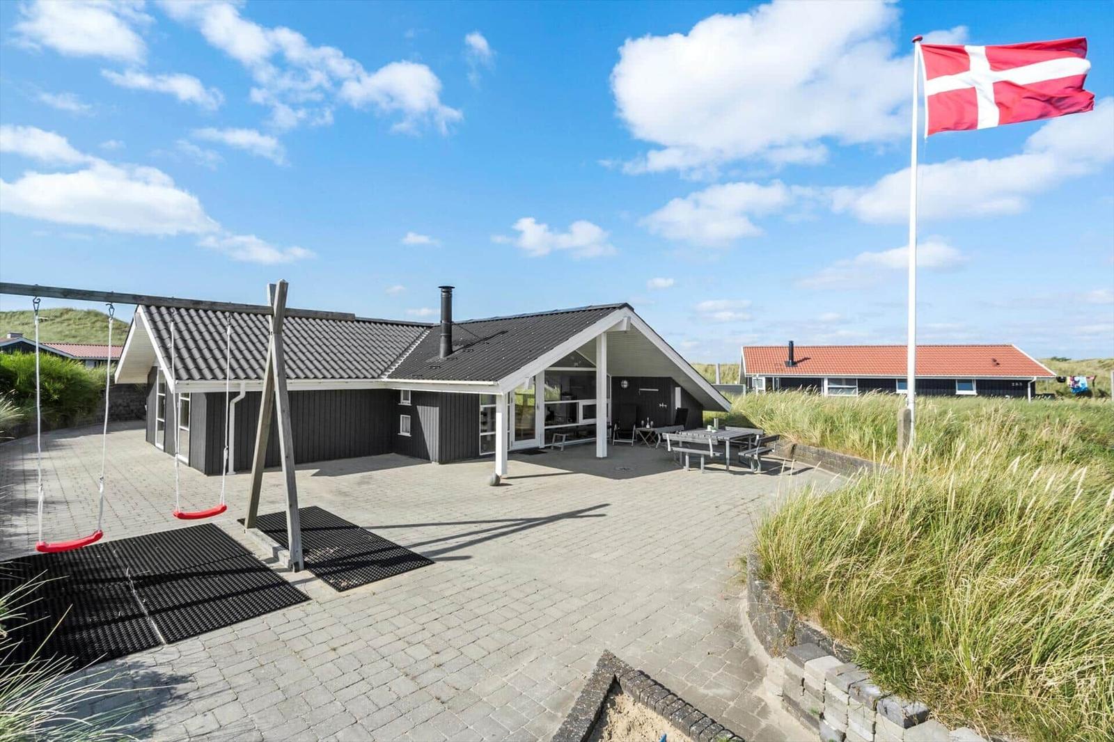 Haus mit Terrasse, Spielplatz und dänischer Flagge vor grüner Vegetation.