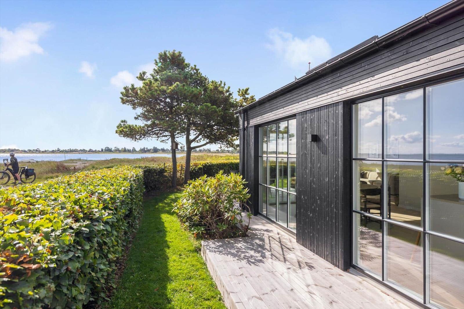 Moderne Villa mit großem Fenster, Blick auf Wasser und Grün. Holzterrasse und Hecke im Vordergrund.