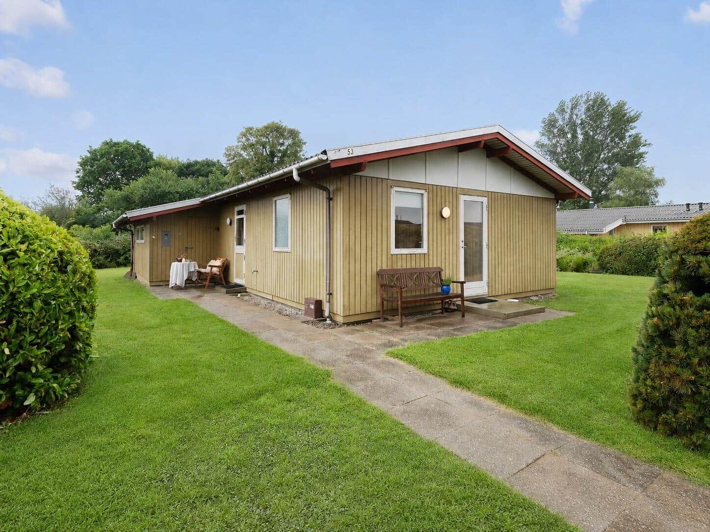 Haus mit Holzfassade, Terrasse und Garten. Holzbank und Tisch stehen draußen.