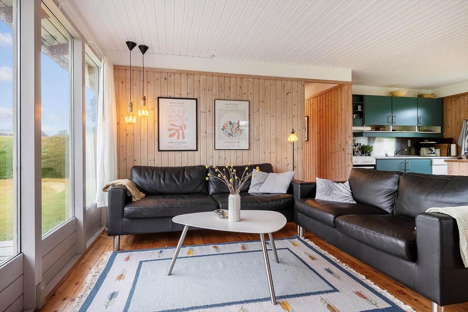 Wohnbereich mit zwei schwarzen Sofas, Holzwand und großem Fenster. Küche im Hintergrund.