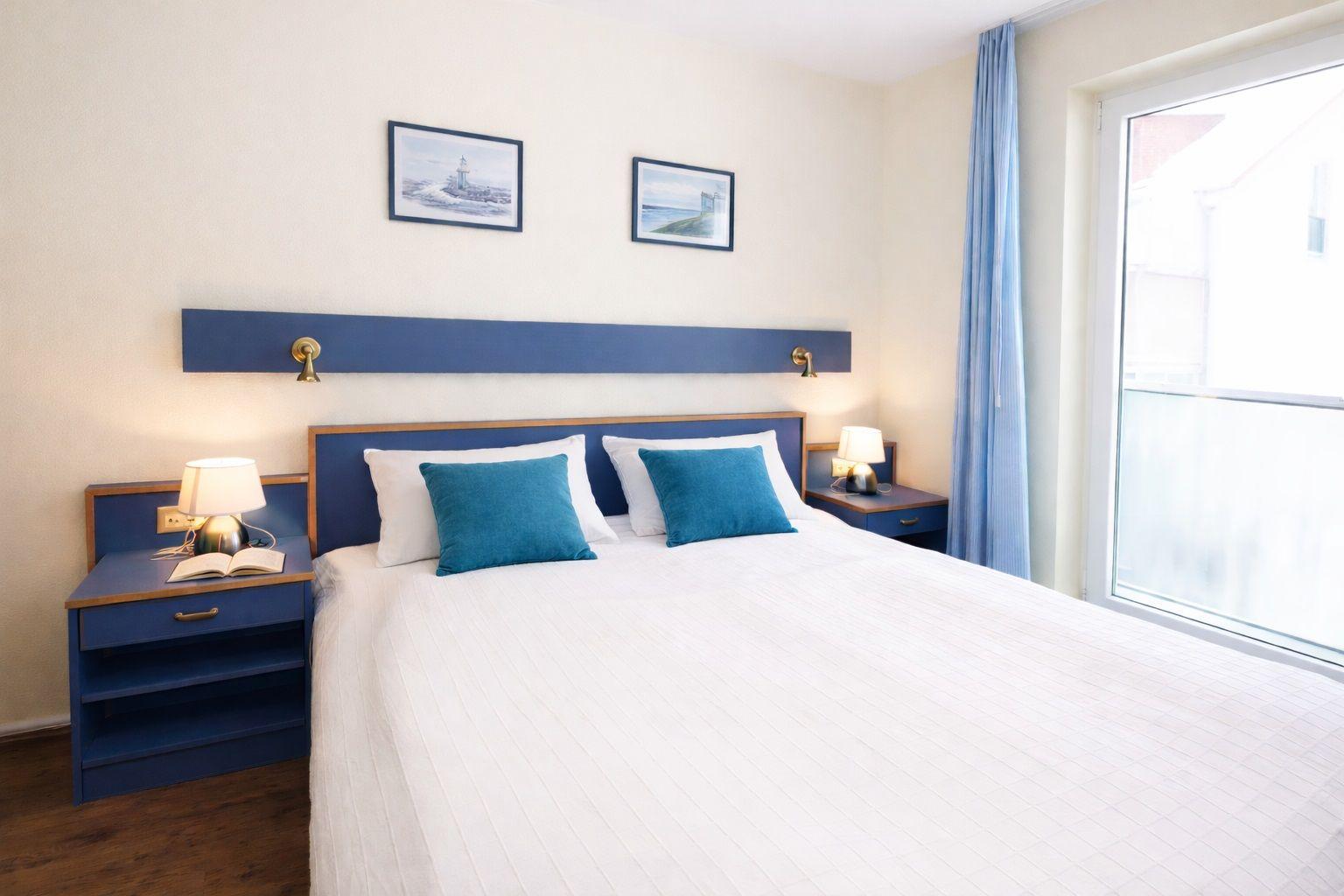 Schlafzimmer mit Bett, Nachttischen und Fenster. Wandbilder und blaue Vorhänge.