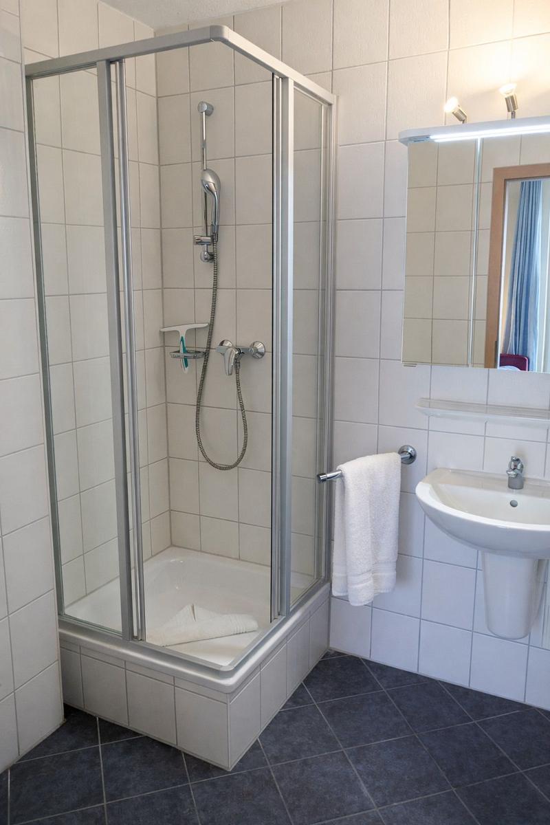 Duschkabine mit Handdusche, Waschbecken und Spiegel. Fliesenboden und weiße Wände.