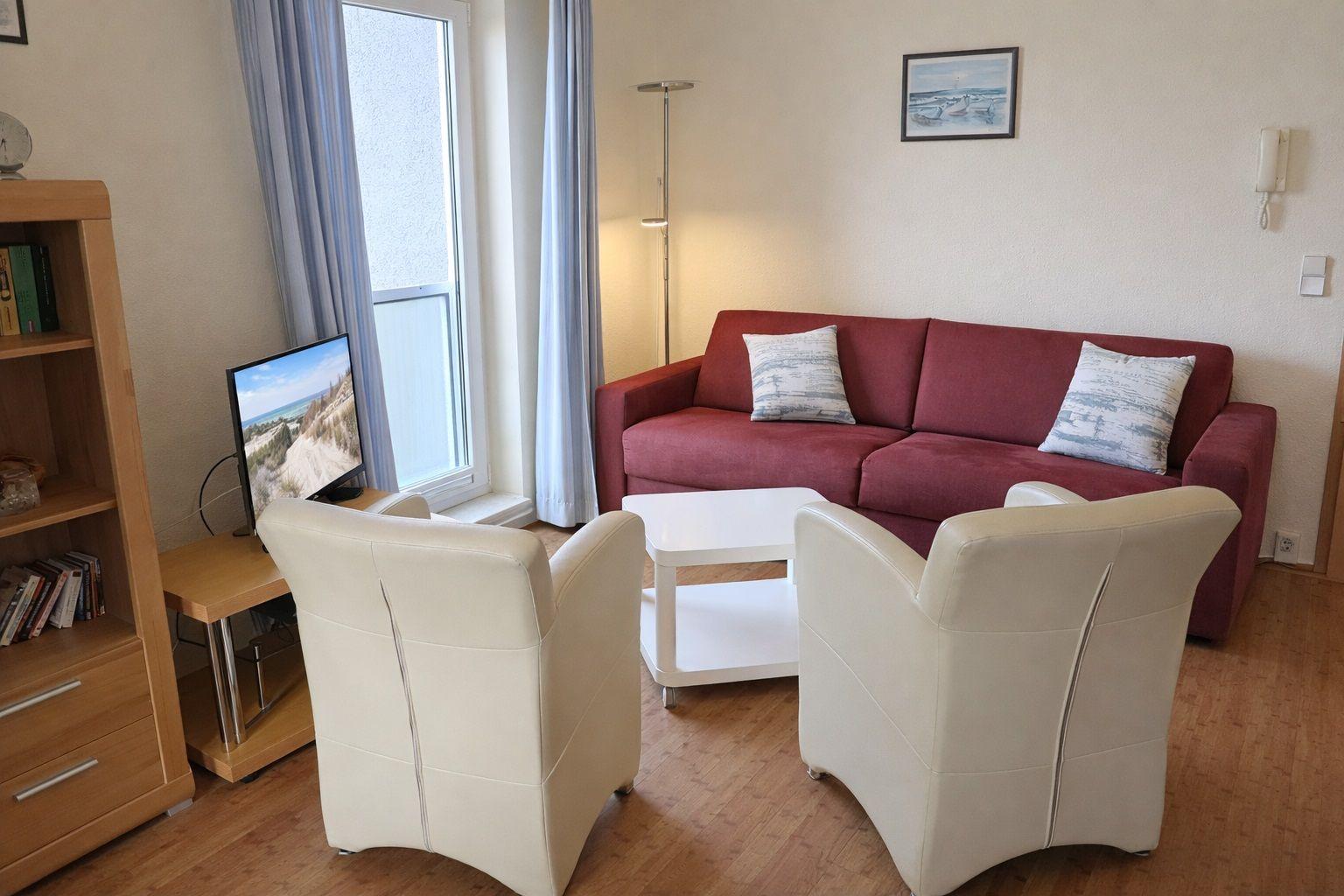 Wohnzimmer mit rotem Sofa, zwei weißen Sesseln und Fernseher an der Wand.
