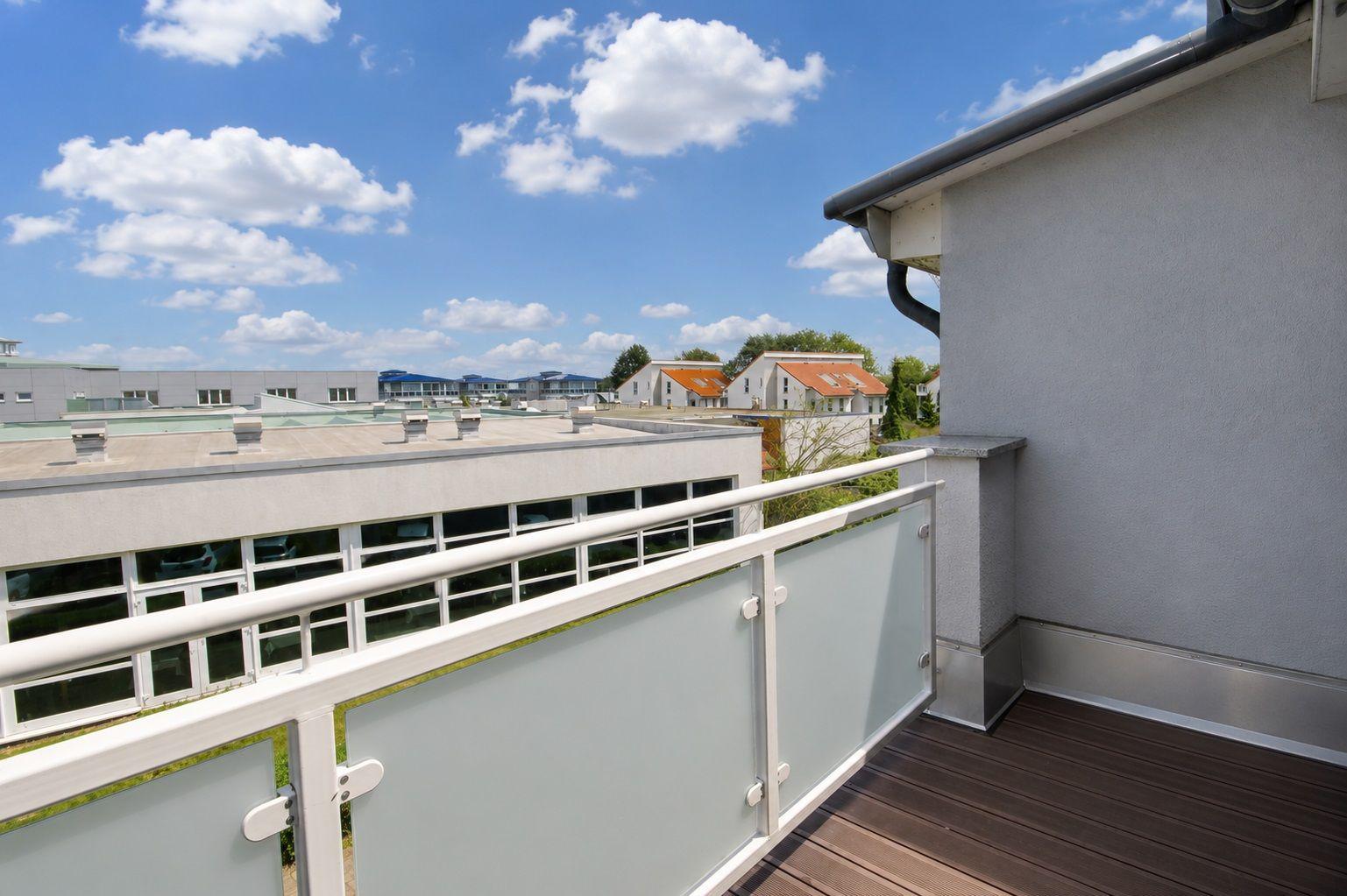 Terrace with wooden floor and glass railing. View of buildings and sky.