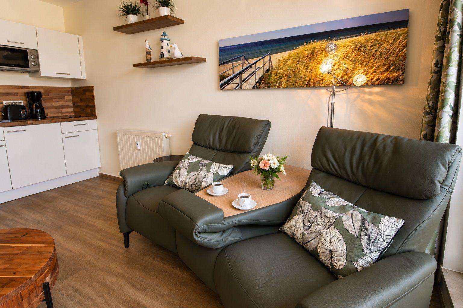Two green recliners with cushions and table. Wall picture and kitchen area visible.