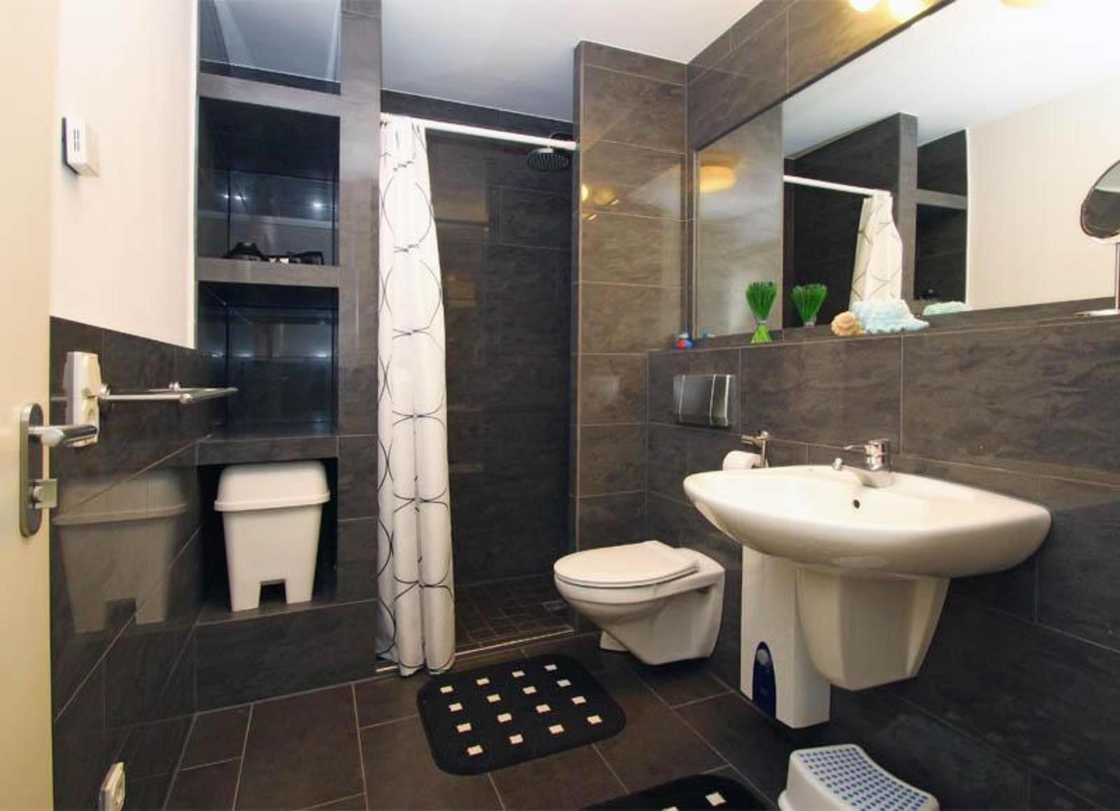 Badezimmer mit Dusche, Waschbecken und Toilette. Dunkle Fliesen und Spiegel.