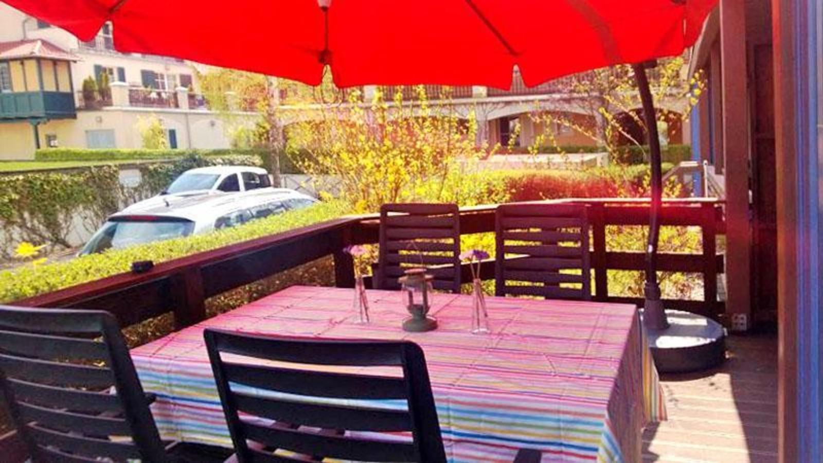 Terrasse mit Tisch, Stühlen und rotem Sonnenschirm. Blick auf grüne Hecke und Parkplatz.