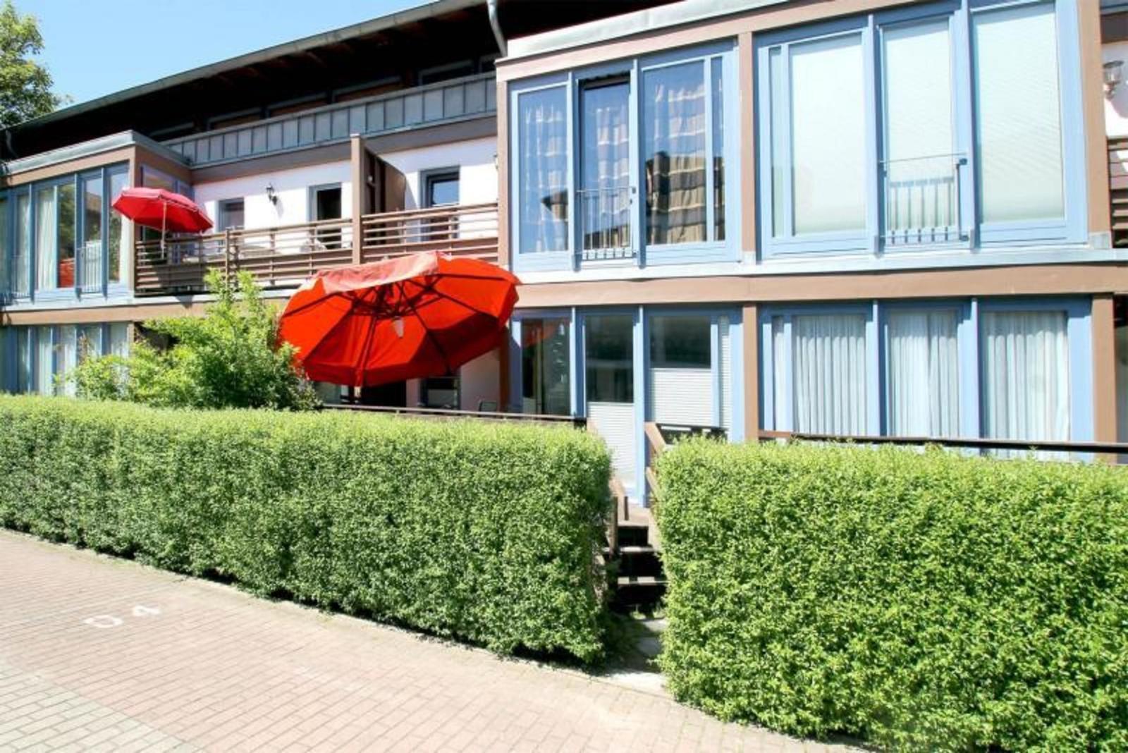 Moderne Ferienwohnung mit Balkon, roten Sonnenschirmen und grüner Hecke.