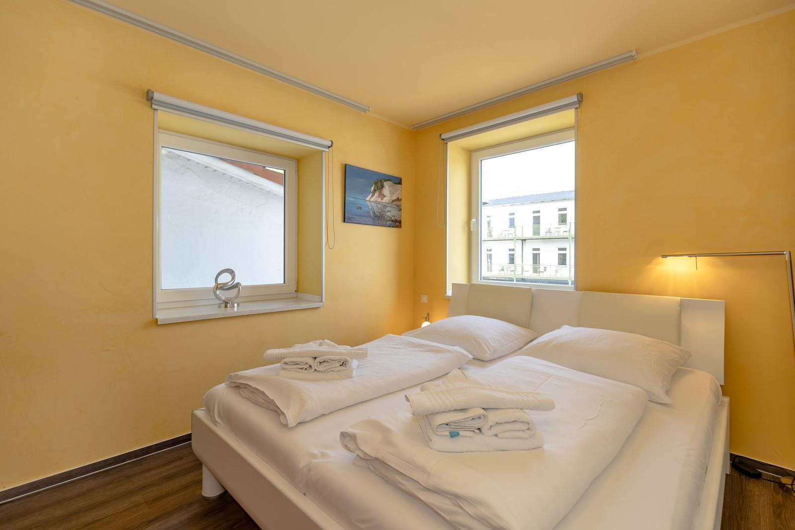 Schlafzimmer mit weißem Bett, gelben Wänden und zwei Fenstern. Auf dem Bett liegen gefaltete Handtücher.