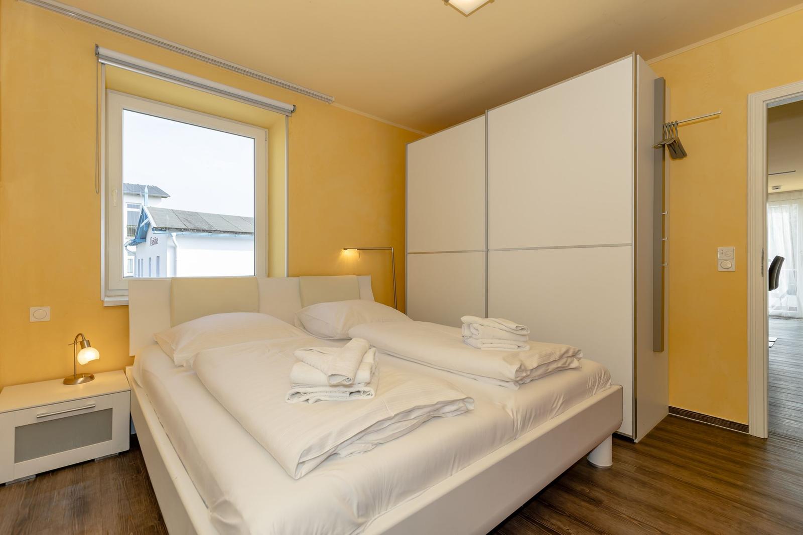 Schlafzimmer mit weißem Bett, Schrank und Fenster. Holzboden und gelbe Wände.