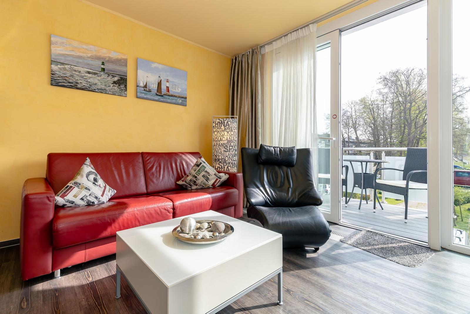 Ein Wohnzimmer mit rotem Sofa, schwarzer Sessel und Balkon. Wandbilder und Couchtisch sind sichtbar.