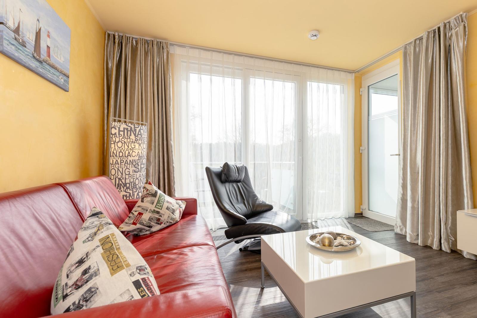 Ein Wohnzimmer mit rotem Sofa, schwarzer Sessel und weißem Couchtisch. Gelbe Wände und große Fenster.