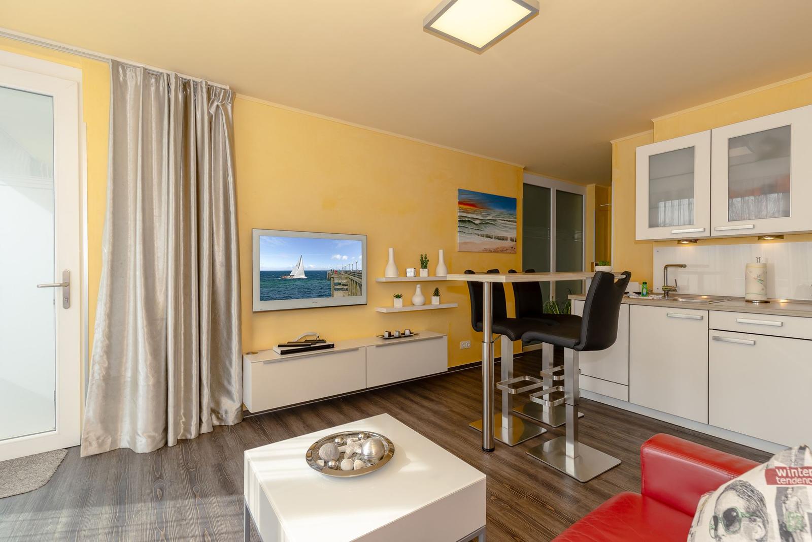 Wohnung mit Küche, Esstisch und Fernseher an gelber Wand.