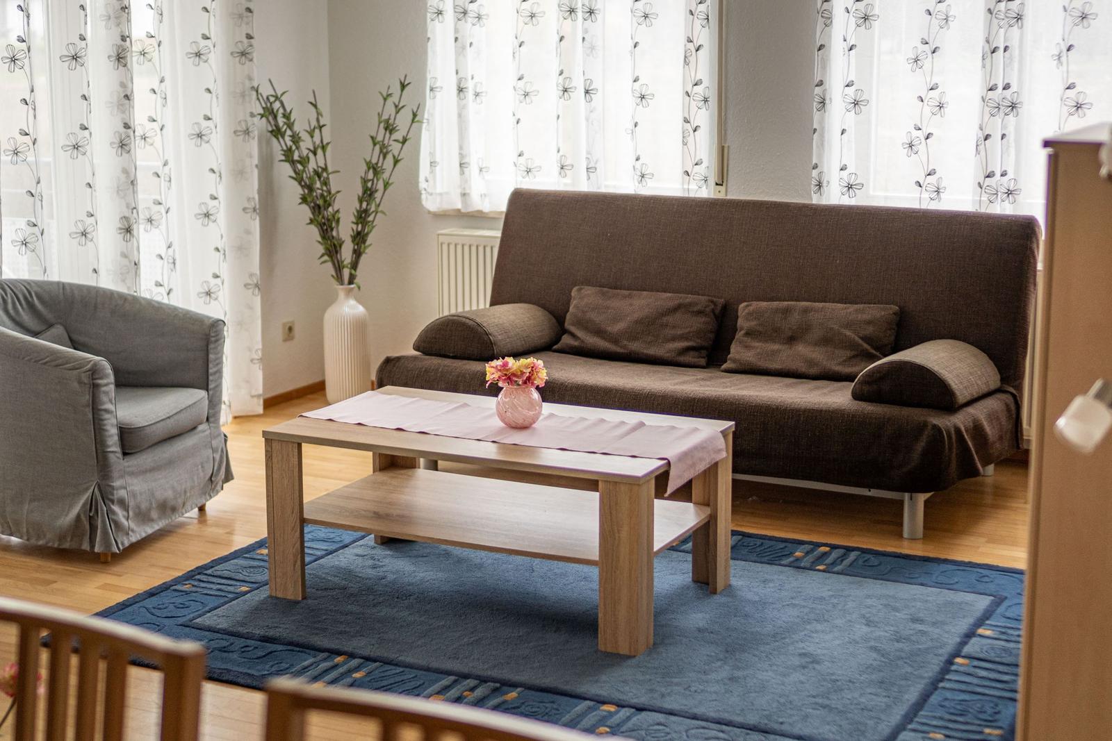 Wohnzimmer mit braunem Sofa, Holztisch und blauem Teppich vor Fenstern mit floralen Vorhängen.