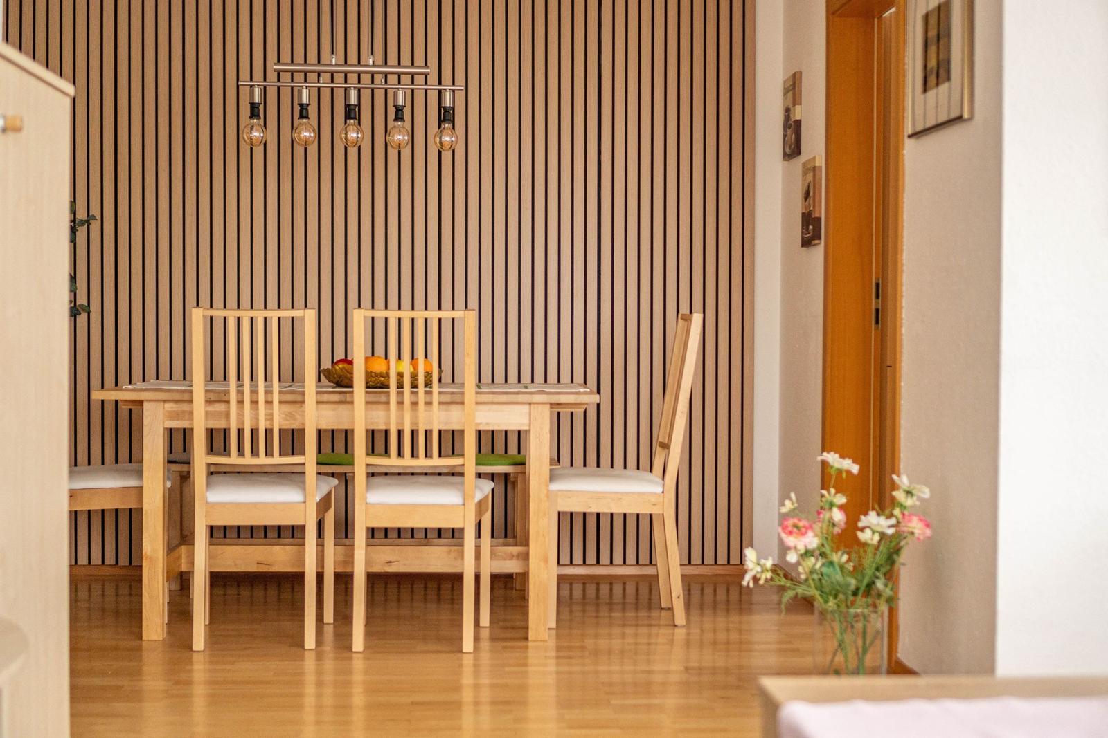 Esszimmer mit Holztisch, vier Stühlen und vertikaler Holzwand. Deckenleuchte mit Glühbirnen.