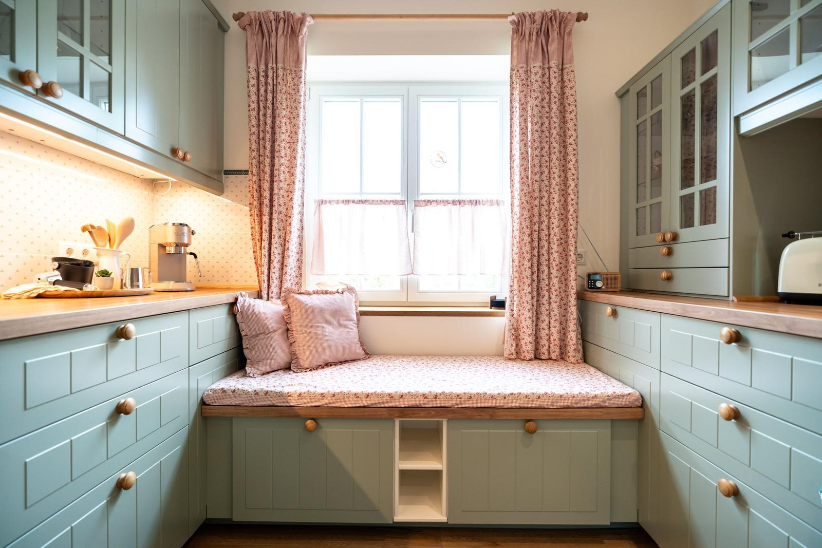 Küche mit Sitzbank unter Fenster, hellblauen Schränken und rosa Vorhängen.