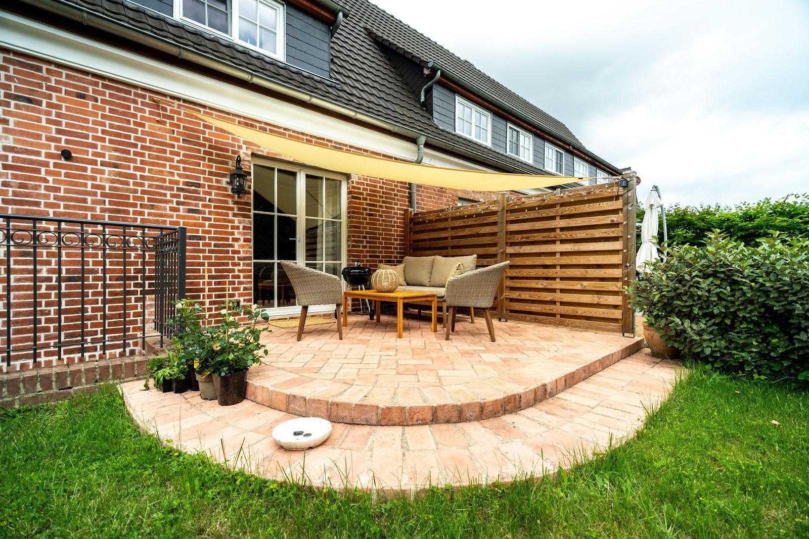 Terrasse mit Sitzgruppe, Sonnensegel und Holzgeländer. Roter Ziegelhof und grüner Rasen.