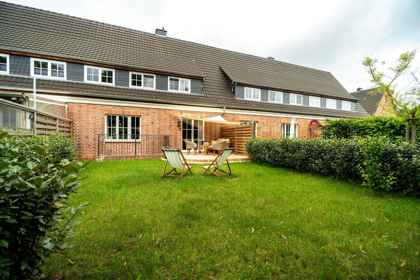 Haus mit Garten, Terrasse und Liegestühlen. Holzgeländer und Hecke umgeben den Außenbereich.