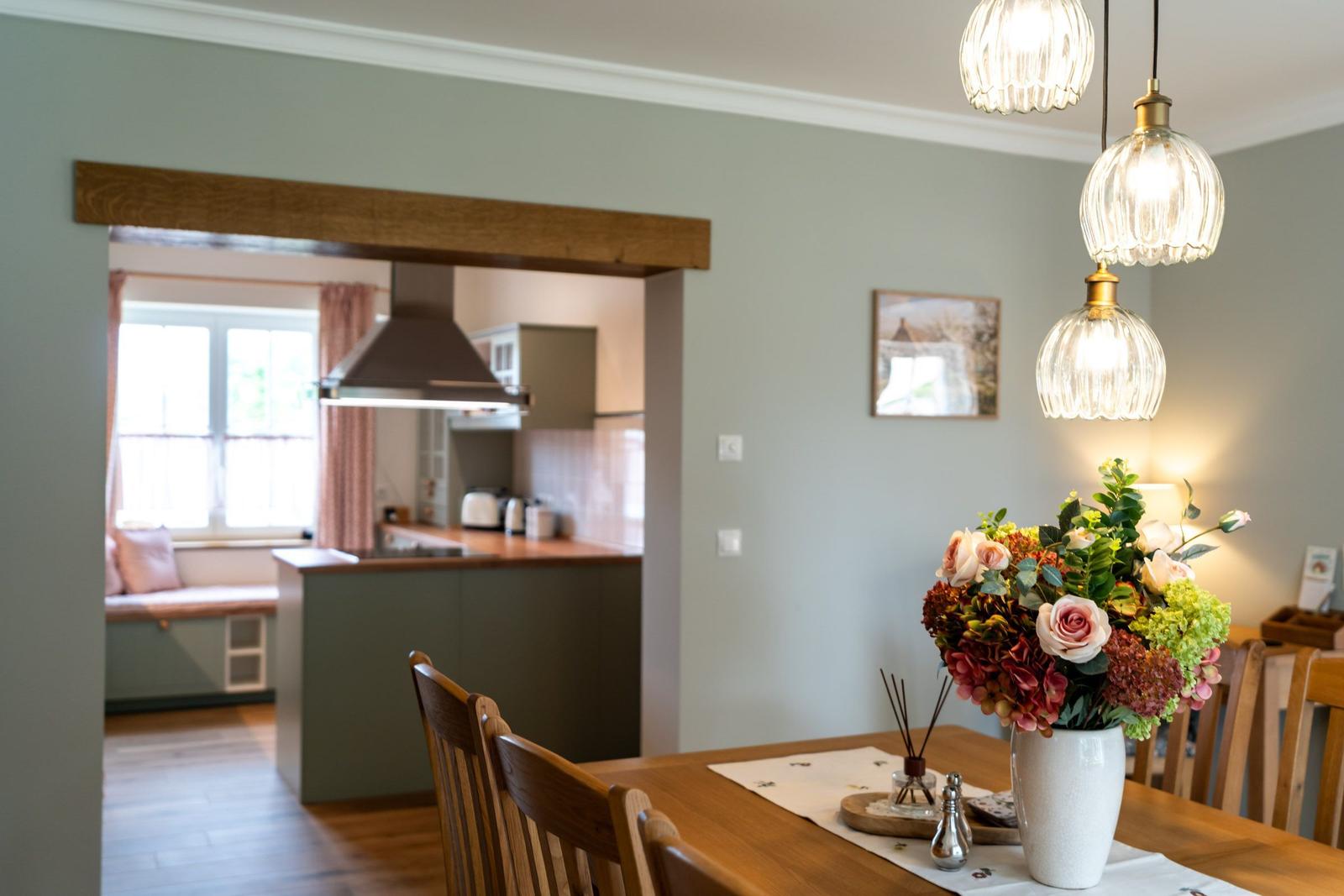 Esszimmer mit Holztisch und Blumenarrangement. Küche im Hintergrund mit Fenster und Sitzbank.