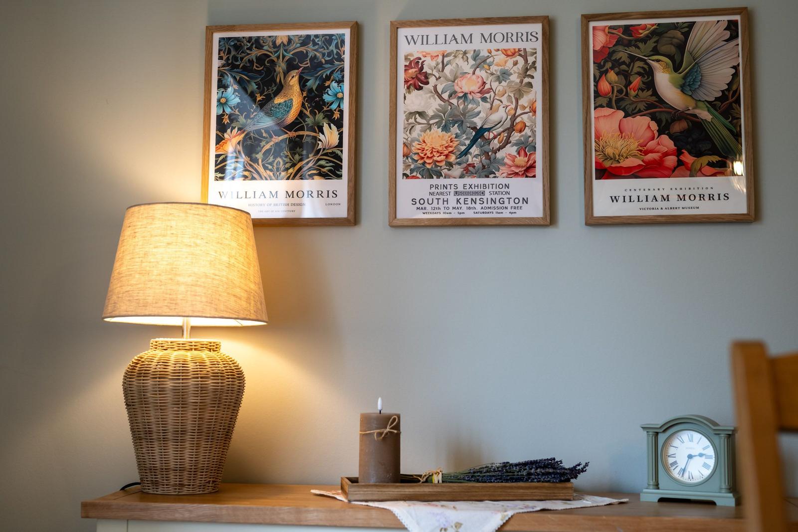 Wand mit drei gerahmten William Morris Poster, Lampe, Kerze und Uhr auf einem Tisch.
