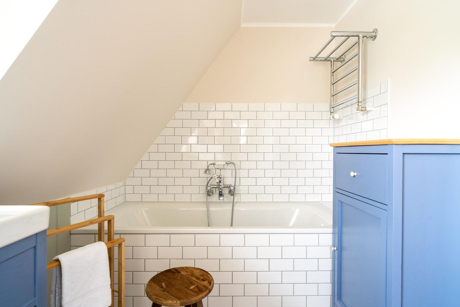 Badezimmer mit Badewanne, weißen Fliesen und blauen Schränken unter schrägem Dach.