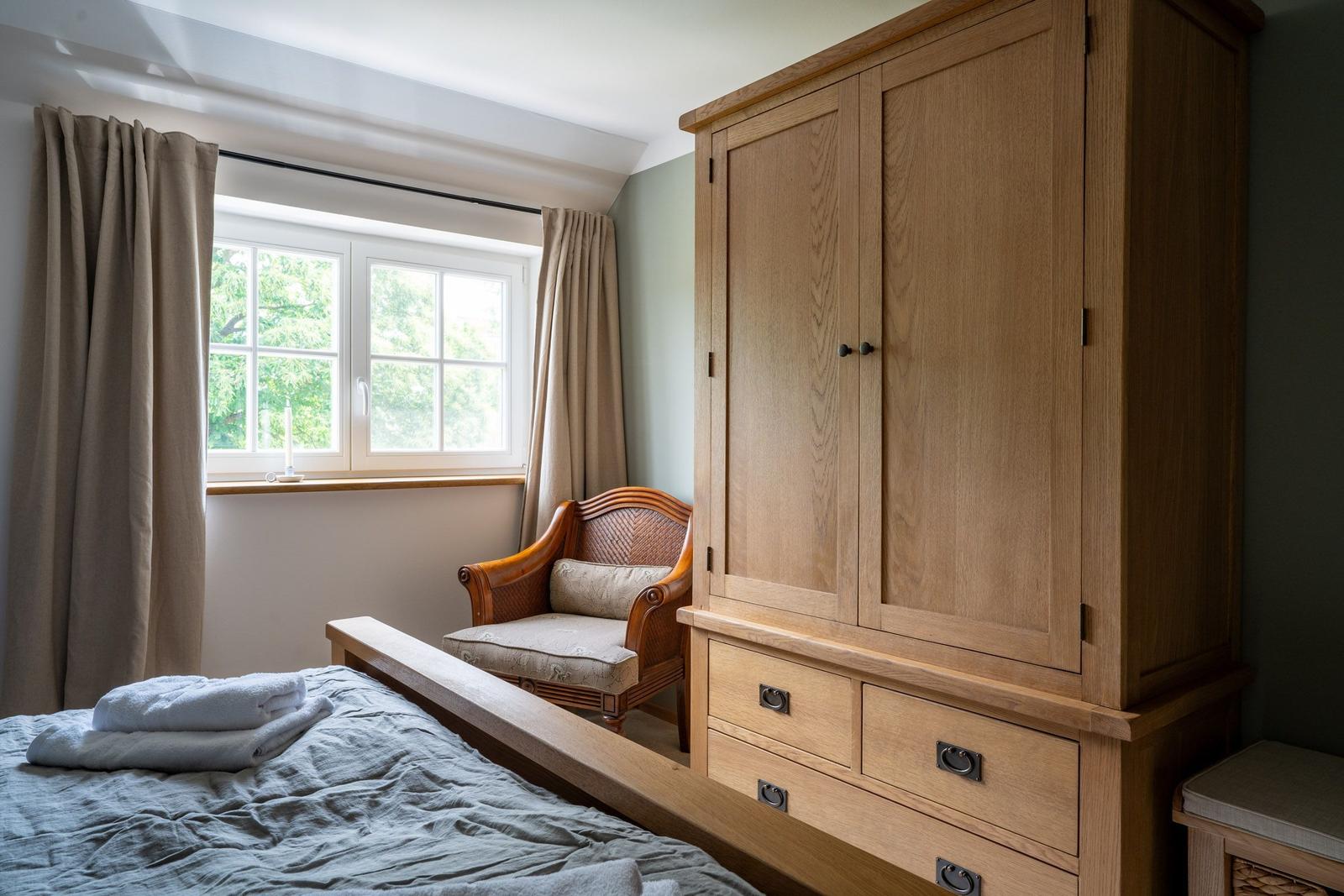 Schlafzimmer mit Holzbett, Kleiderschrank und Sessel vor Fenster mit Blick auf Bäume.