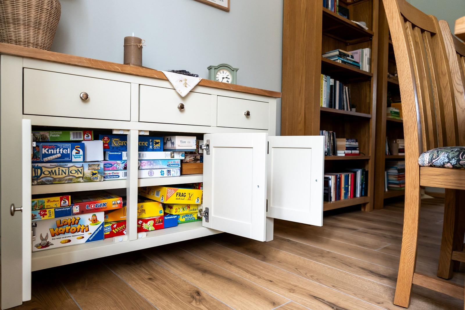 Weißer Schrank mit offenen Türen zeigt Brettspiele. Holzregal und Stuhl im Hintergrund.