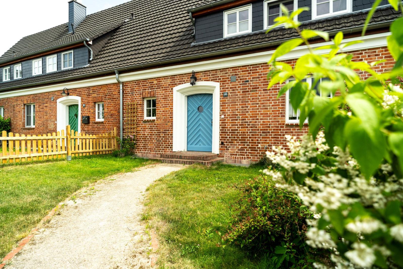 Rotes Backsteingebäude mit blauer Tür und grünem Tor. Gartenweg und Blumen im Vordergrund.