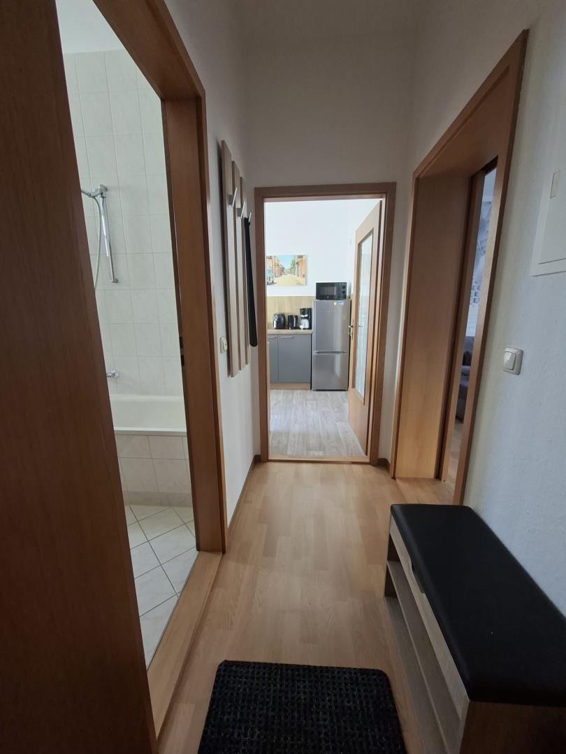 Hallway with view into kitchen, bathroom, and bedroom. Wooden floor and black bench.