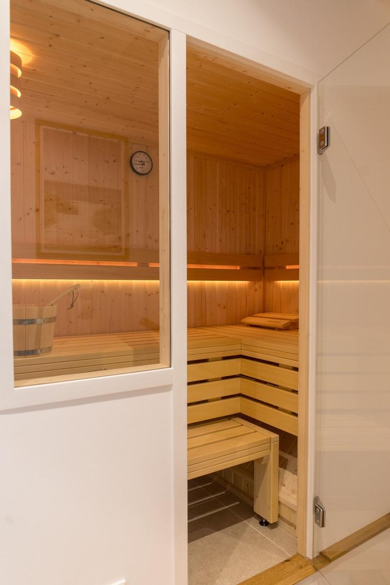 Sauna mit Holzbank und Eimer. Wanduhr und Beleuchtung sichtbar.