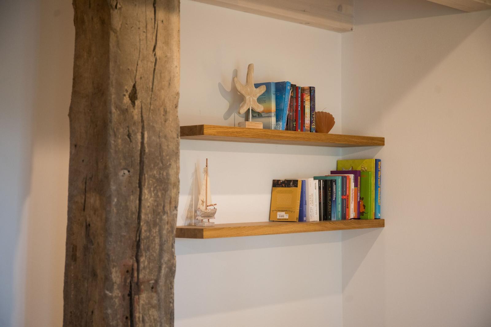 Zwei Holzregale an der Wand mit Büchern und Meeressymbolen. Links Holzpfosten.