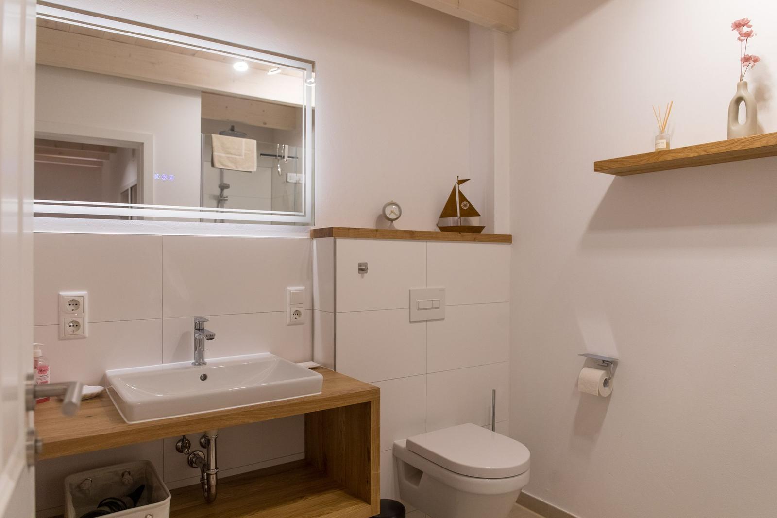 Badezimmer mit Waschbecken, Spiegel und Toilette. Holzregal und Dekoration.