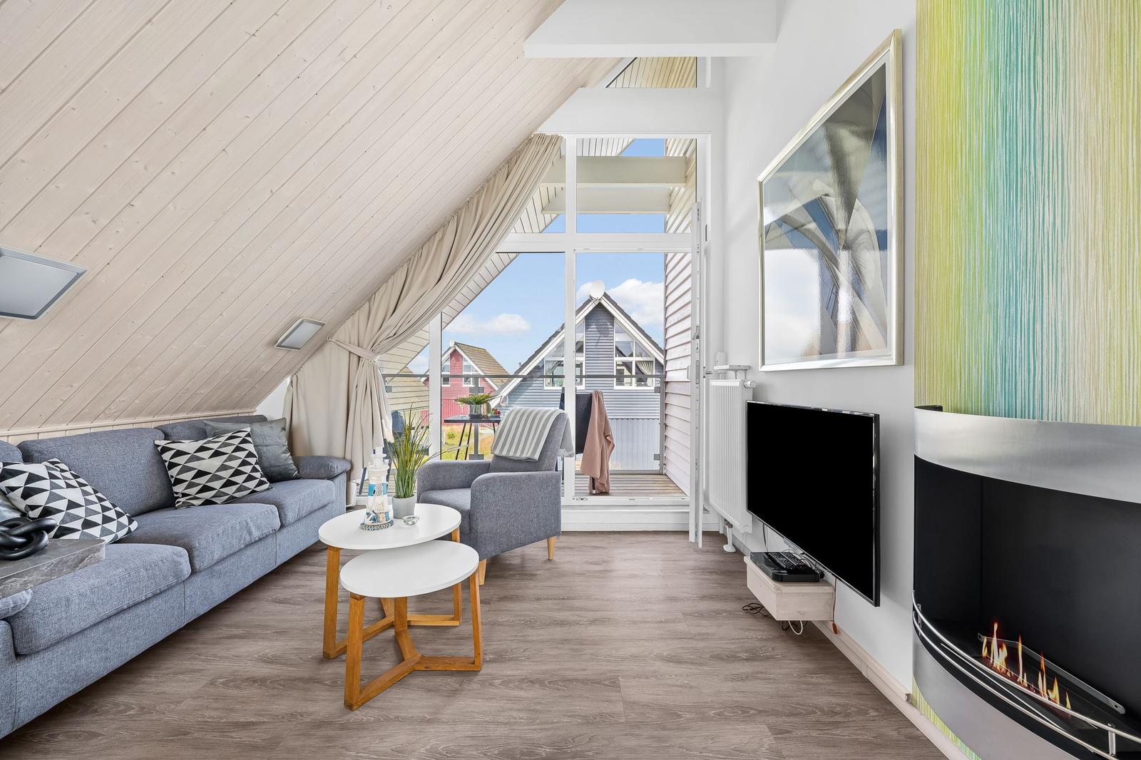 Wohnzimmer mit Sofa, Sessel, Fernseher und Kamin. Blick auf Balkon und Nachbarhäuser.