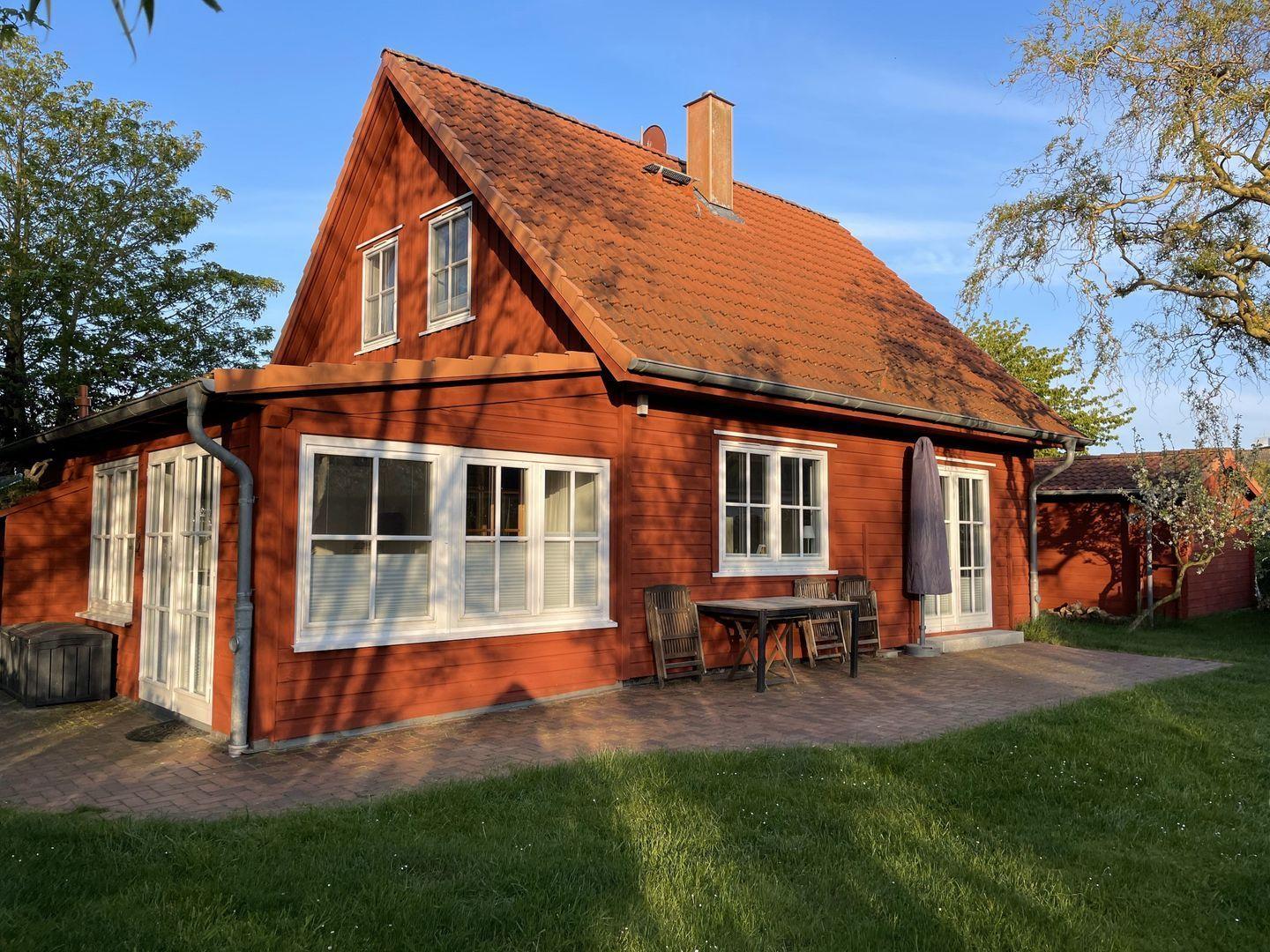 Rotes Holzhaus mit Terrasse, Tisch und Stühlen. Grünfläche und Bäume im Hintergrund.