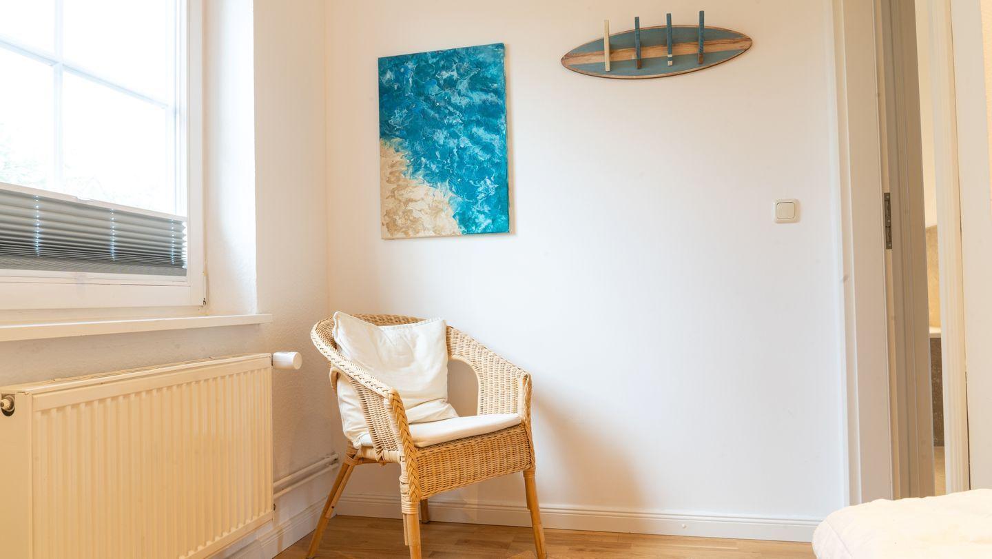 Ein Raum mit Stuhl, Wandbild und Fenster. Ein Bett ist teilweise sichtbar.