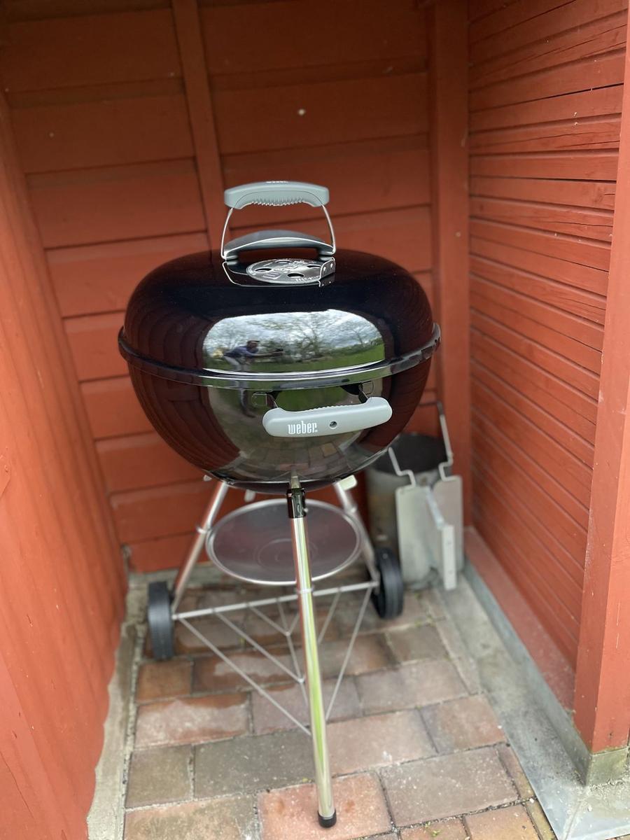 Schwarzer Grill mit silbernen Griffen auf einem Metallgestell vor roten Holzwänden.