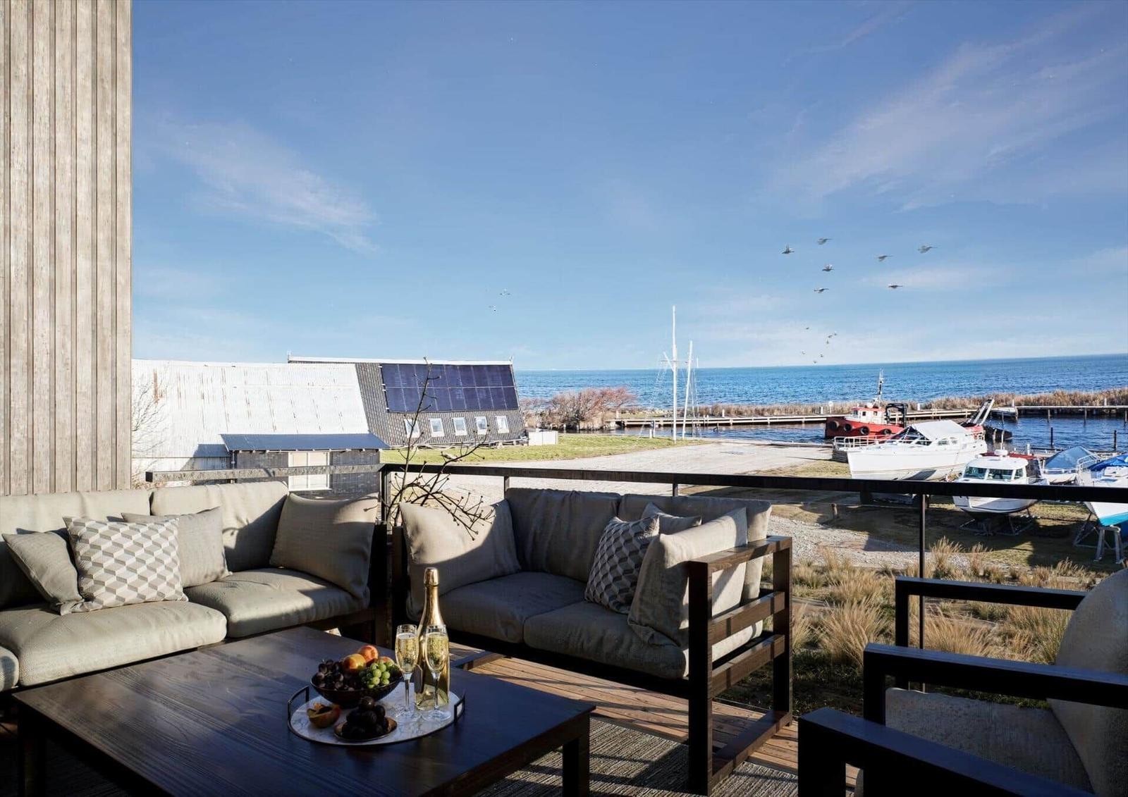 Terrasse mit Sofa, Tisch und Meerblick. Boote liegen am Hafen. Himmel klar, Vögel fliegen.