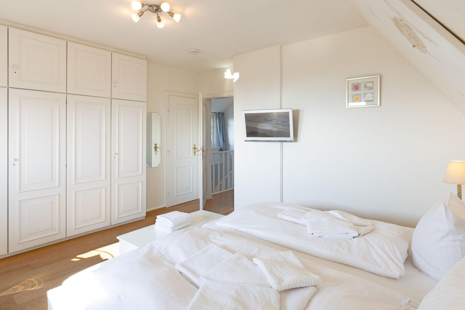 Schlafzimmer mit weißem Bett, Kleiderschrank und Wandfernseher.