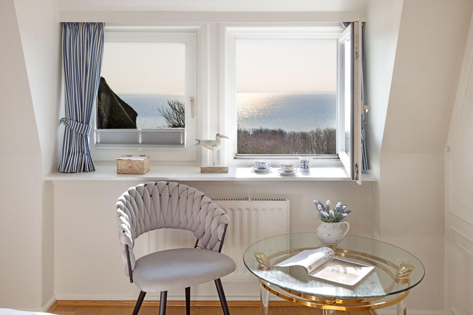 Ein Raum mit Blick aufs Meer, zwei Fenster, Stuhl und Glas-Tisch.