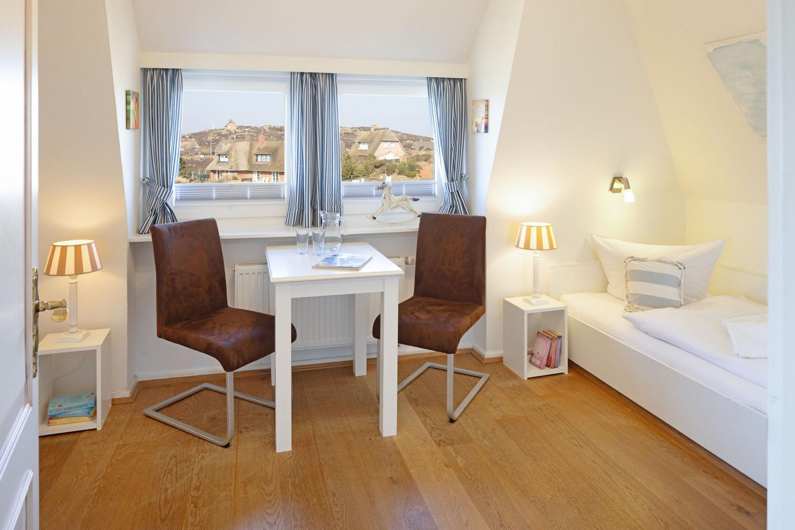 Zimmer mit Schlafcouch, Tisch und zwei Stühlen. Blick aus dem Fenster auf Dächer.
