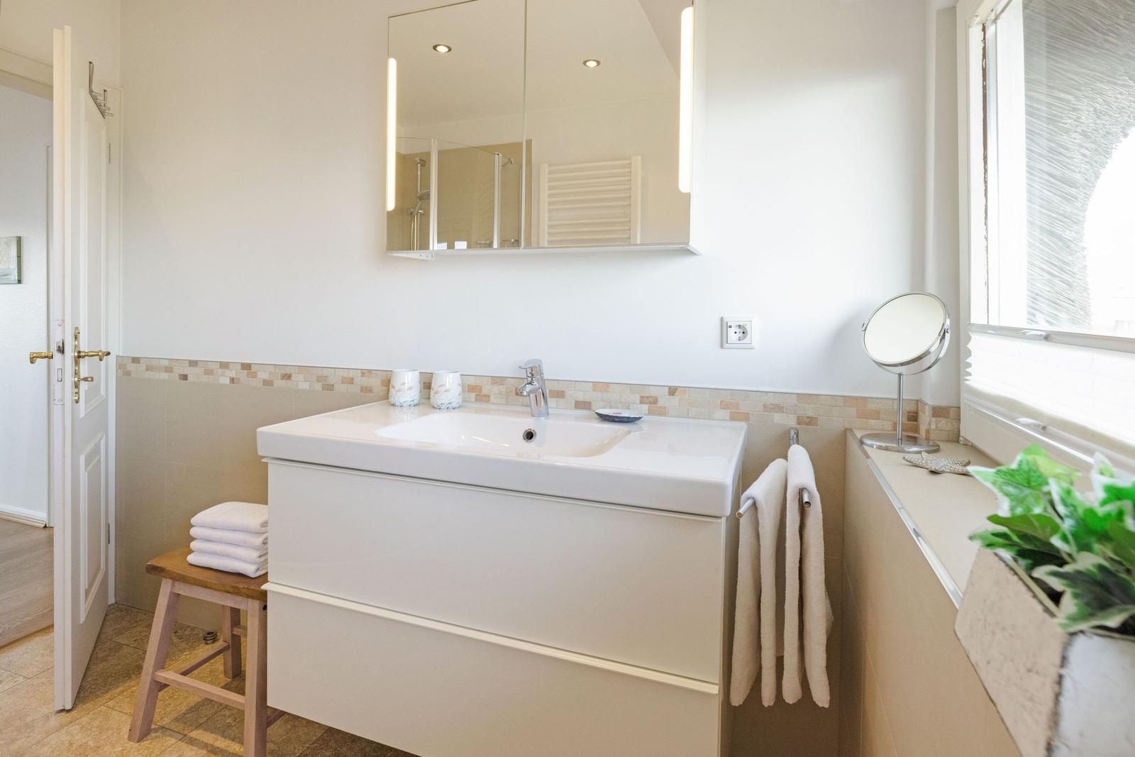 Badezimmer mit Waschbecken, Spiegel, Handtuchhalter und Pflanze an Fensterbank.
