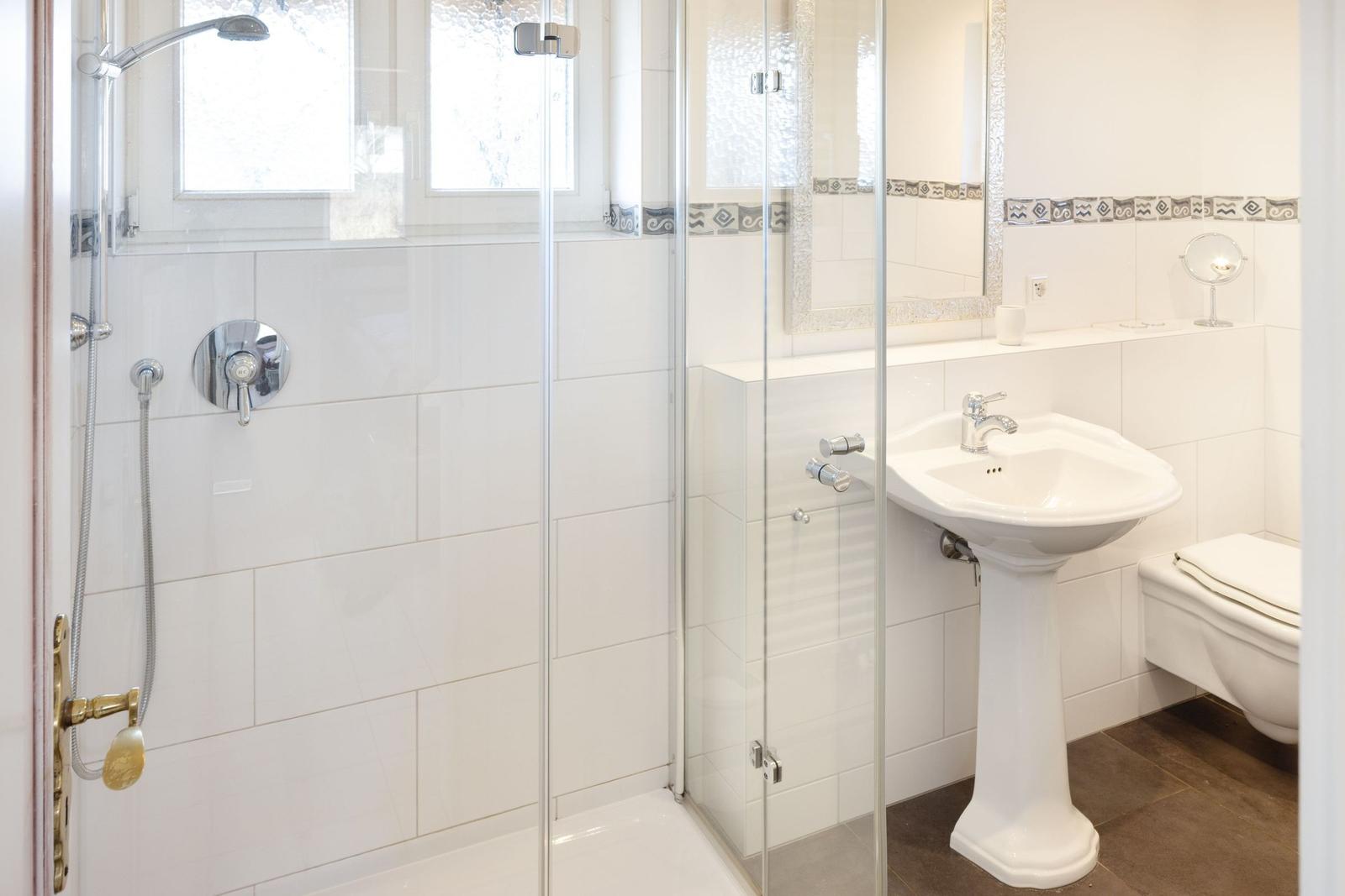 Badezimmer mit Dusche, Standwaschbecken und Toilette. Weiße Fliesen und dunkler Boden.