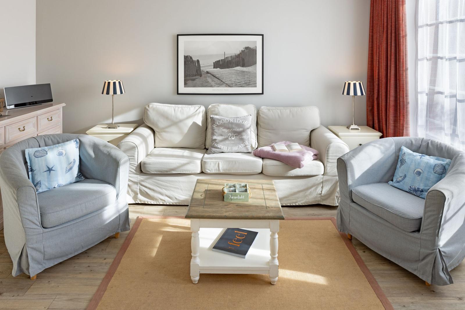 Wohnzimmer mit Sofa, zwei Sesseln, Couchtisch und Fenster mit Vorhängen.