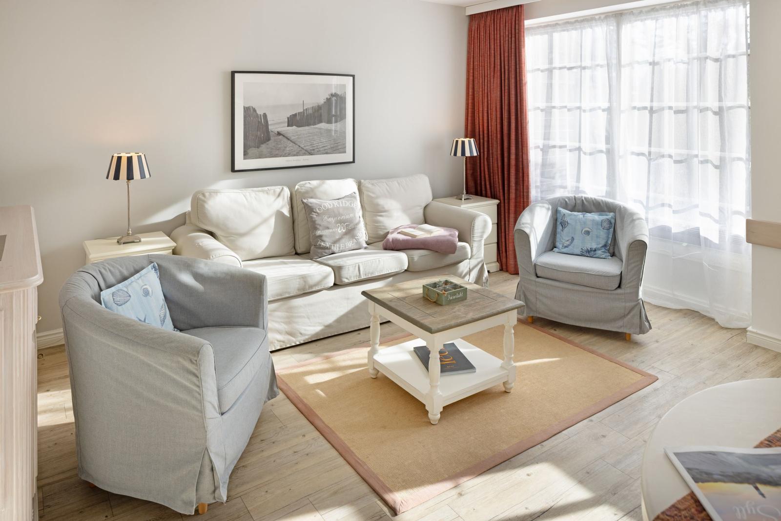 Wohnzimmer mit Sofa, zwei Sesseln, Couchtisch und Fenster mit Vorhängen.